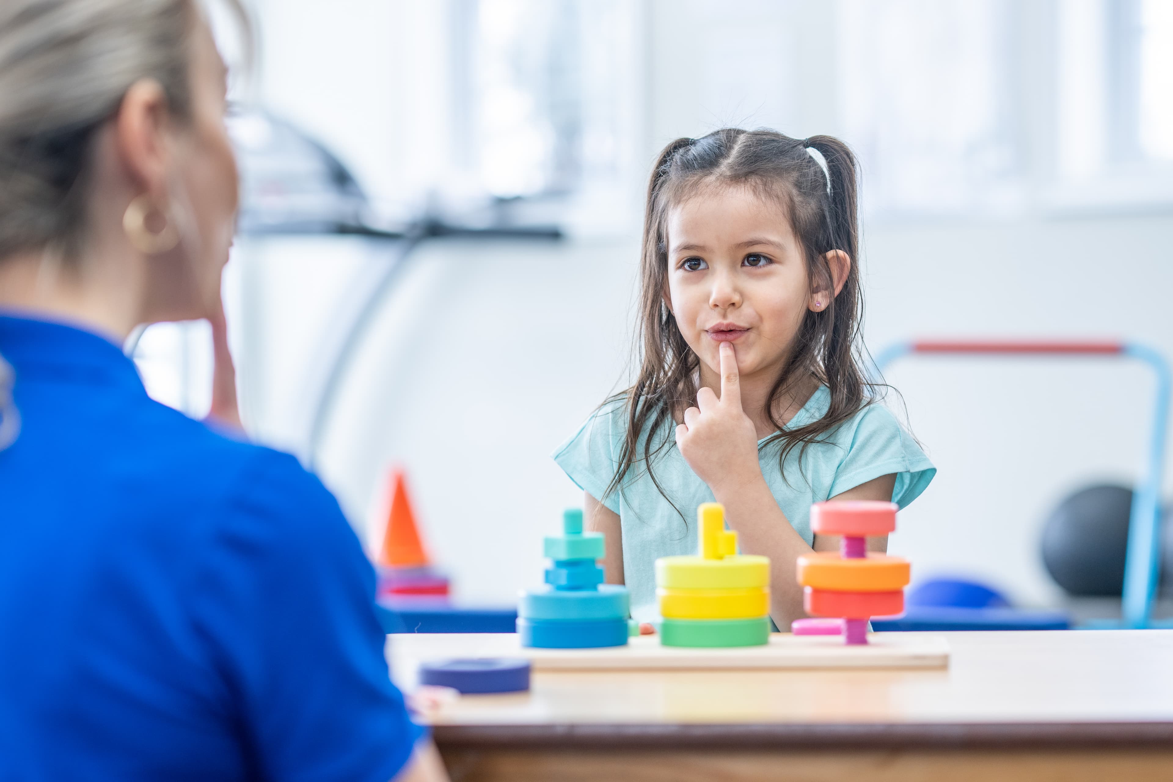 Young girl touches her lip during a speech therapy session with colorful stacking toys.